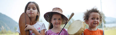 Three young children stand outside. The girl on the left holds a paddle upright. The girl in the middle holds a drum. The boy on the right smiles at the camera.