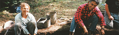 Two people sit within a sectioned archaeological area, using shovels to excavate the earth.