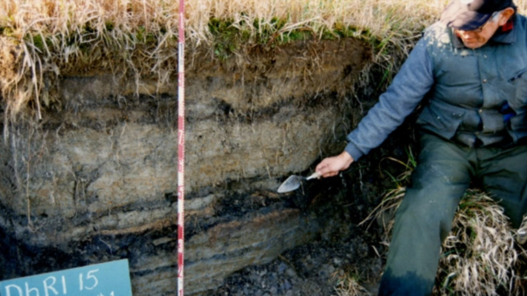 A man leans over an archeological mound, using a shovel to point out the different layers of material.