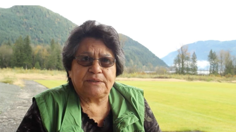 An elder talks to the video camera outside. There are mountains and trees in the background.