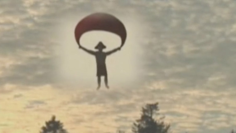 A figure with a cedar-root hat descends through the sky holding a parachute.