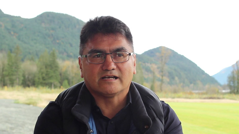 A man sits in front of a soccer field, talking to a video camera.