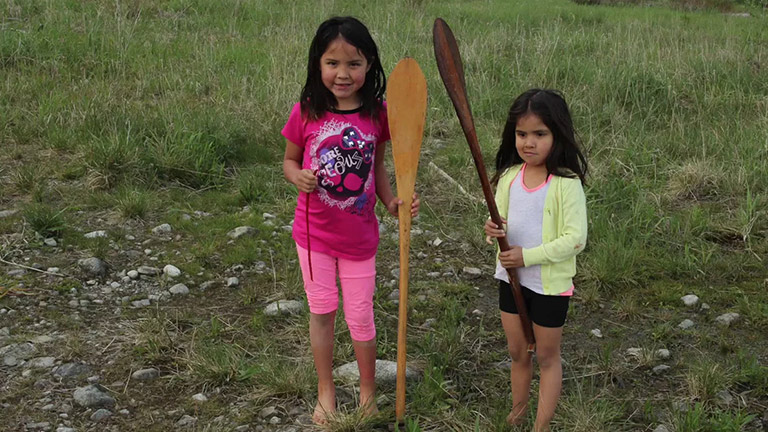 Two young girls pose on the grass with their canoe paddles. 