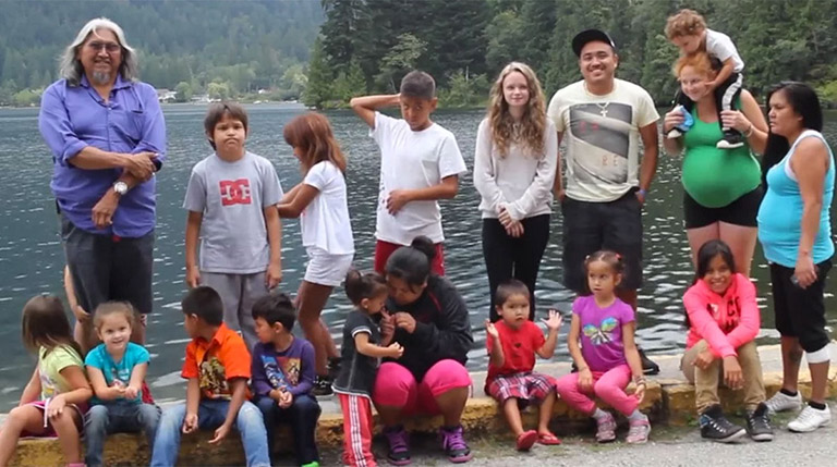 A group of children and young adults pose with their elder for a photograph on the shore of the river. There are mountains and trees in the background.