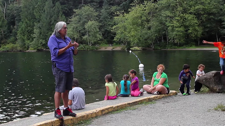 Un aîné raconte une histoire aux enfants et aux membres de la communauté près de la rive.