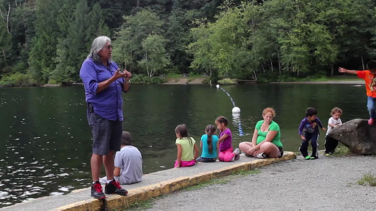 An elder shares a story with children and community members near the shore of the river. 