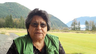 An elder talks to the video camera outside. There are mountains and trees in the background.