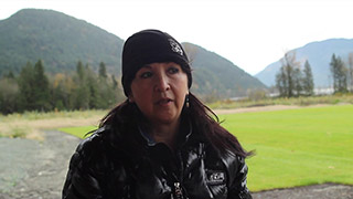 A woman shares her story with a video camera. She sits outside with the mountains and trees behind her.