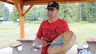 A man stands outdoors playing a hand drum. The drum is decorated with the image of a blue hummingbird. 