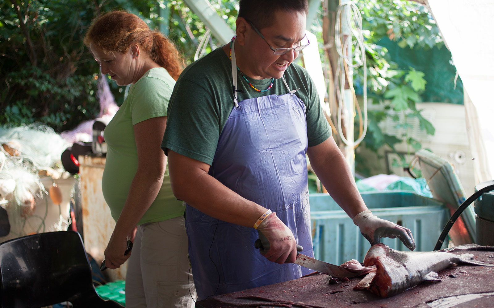 A man is cutting a fish on an outdoor table. He holds the body of the fish with his left hand, and a knife in the other.