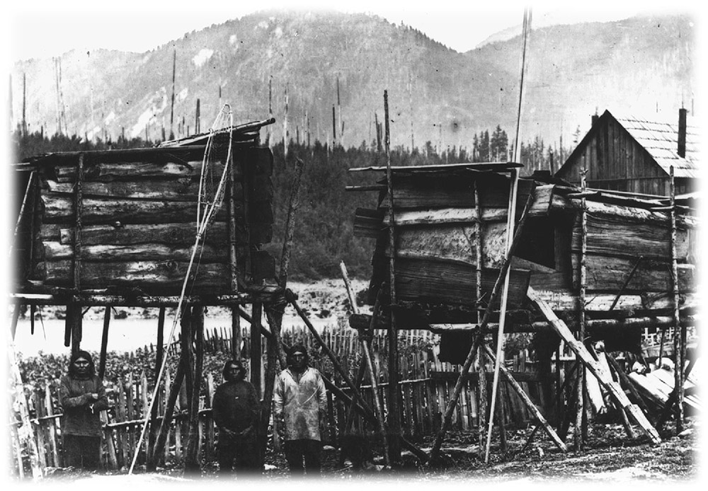 A black and white photograph of raised storage boxes on wooden stilts. There are several men standing in the foreground, and snow-capped mountains in the background.