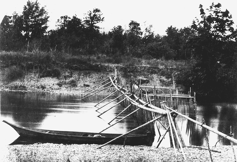Une photo en noir et blanc d’un barrage à poissons dans un canal fluvial. Au premier plan, un canot des Salish de la côte.