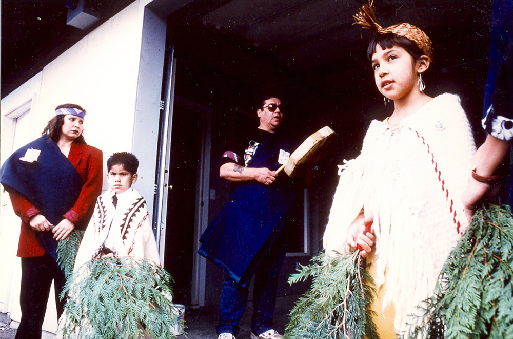 There are two young children dressed in regalia, holding onto cedar branches. A man and woman stand close by.