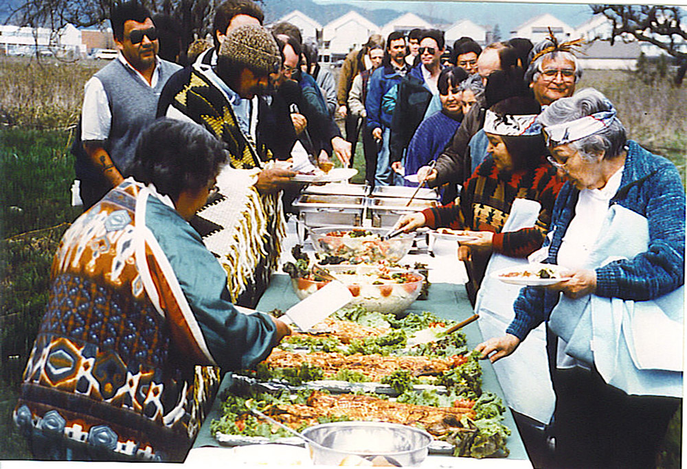 A large group of people are gathered outside, lined up around a long table that is covered in many dishes of food at a First Salmon Ceremony.
