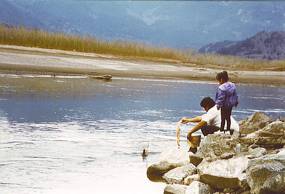 A man and young girl are gathered by the edge of the river; the man is crouching down with an object in his hand, while the girl stands beside him looking onwards.