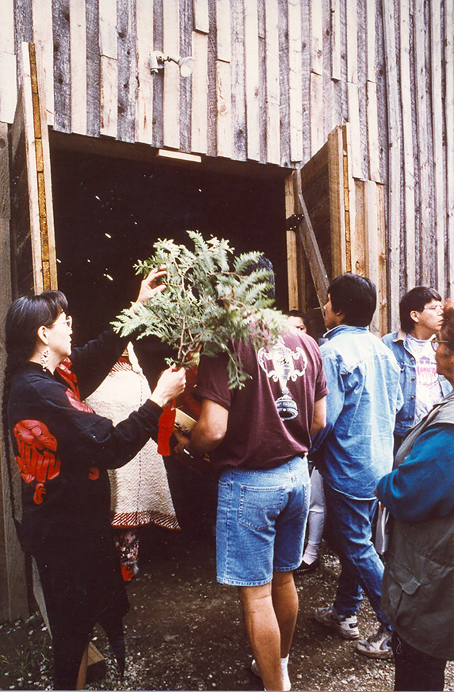 A group of people are gathered outside of a wooden building; they are being brushed with a cedar branch before entering the building.