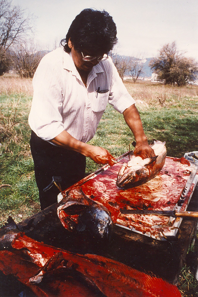 A man stands over a table set up outside; he is preparing salmon for cooking. 