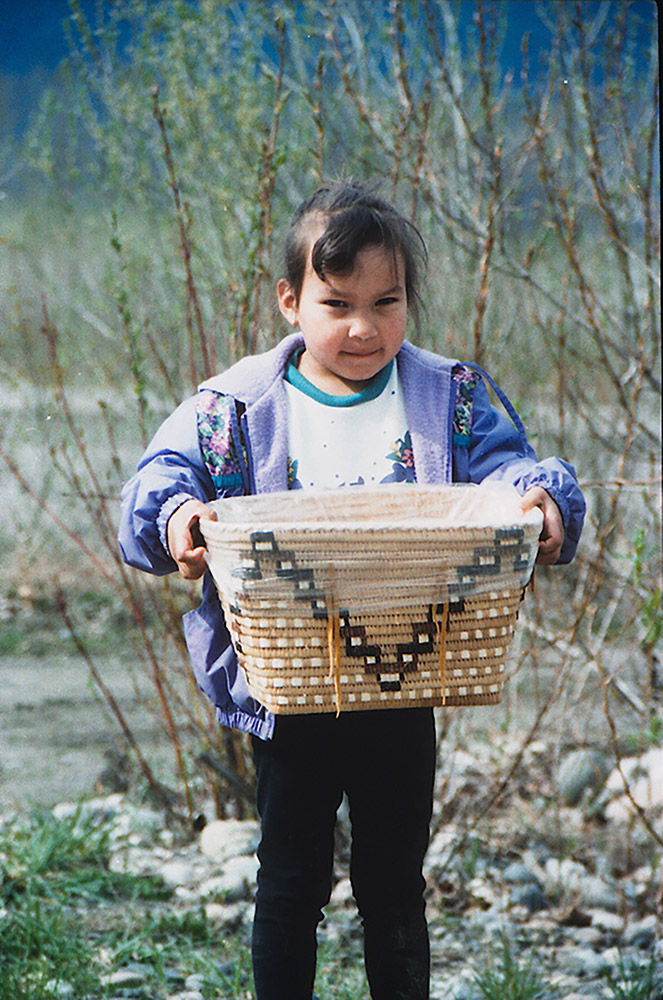 A young girl stands outside, holding onto a medium-sized woven basket with both hands. The basket has a design in the shape of a “V” on the front.