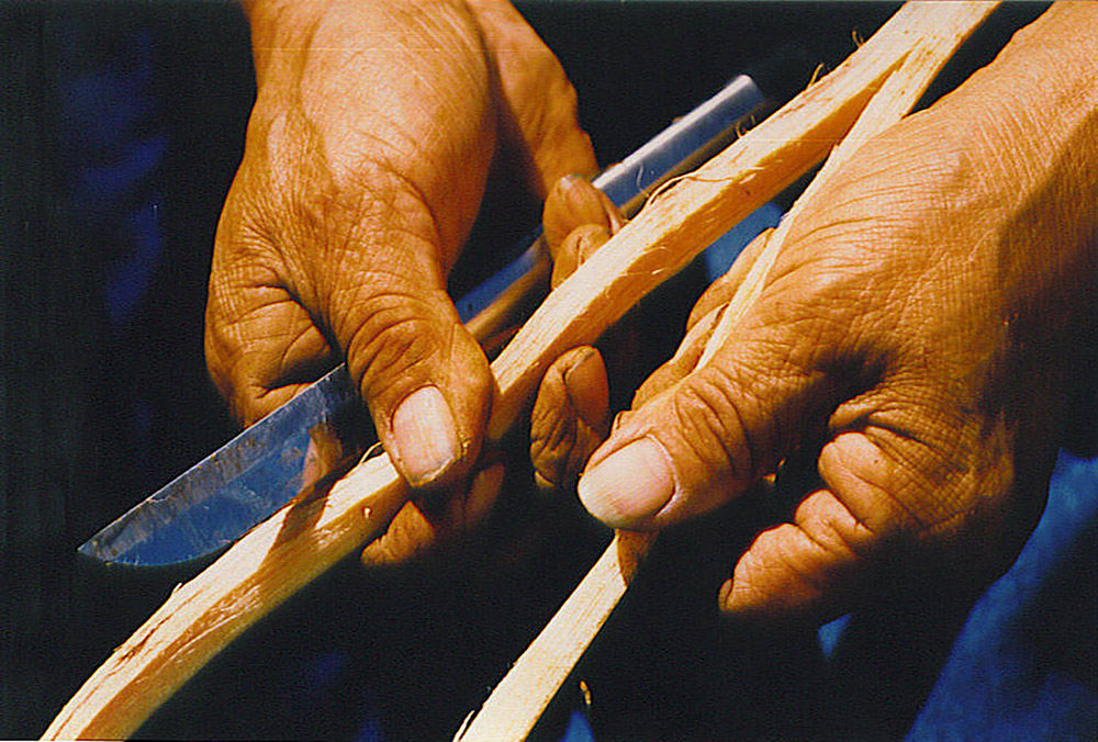 A man uses a knife to separate a cedar root into two halves, still attached at the base.