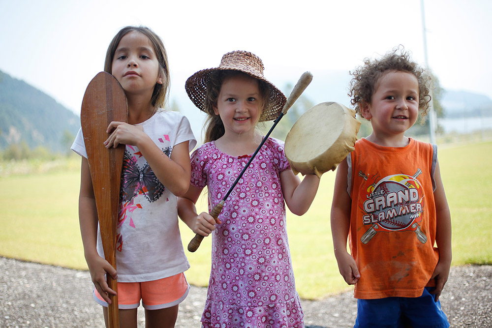 Three young children stand outside. The girl on the left holds a paddle upright, touching her chin. The girl in the middle holds a drum. The boy on the right smiles at the camera.