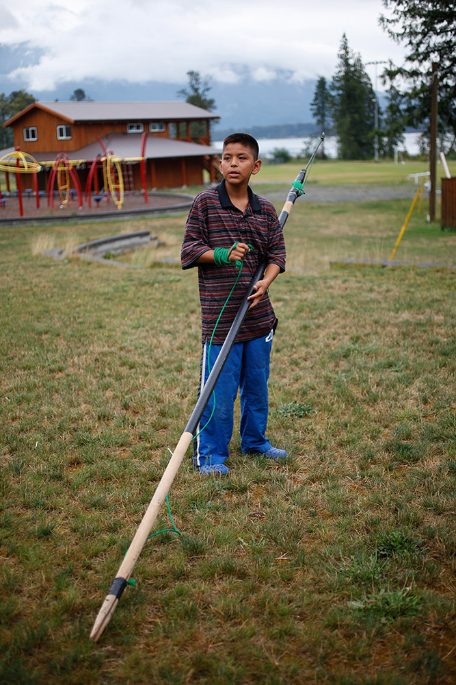 A young boy stands on the grass, holding his homemade fishing spear.