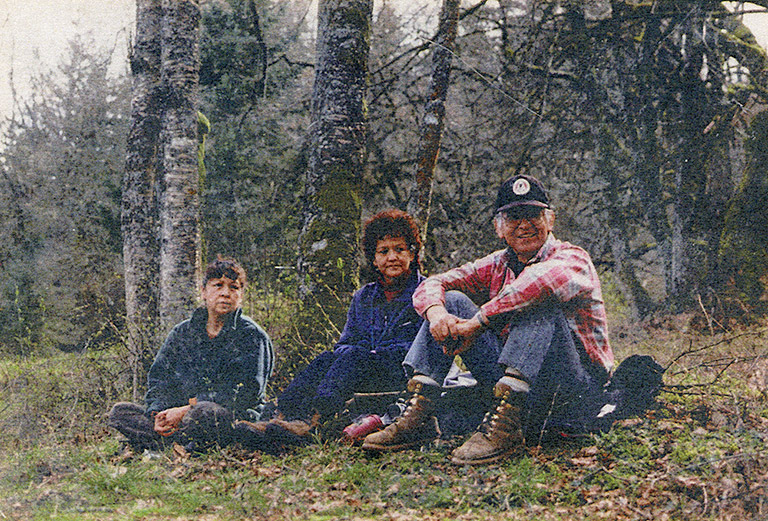 Three adults are sitting on the grass outside, with trees behind them.