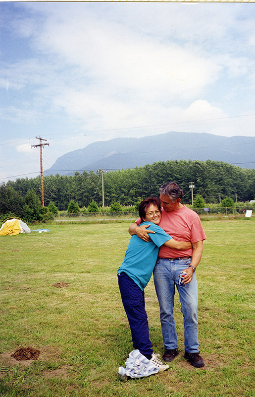 A woman and man stand outside on the grass. She has her arms wrapped around the his waist, and is facing the camera. He is looking at her.