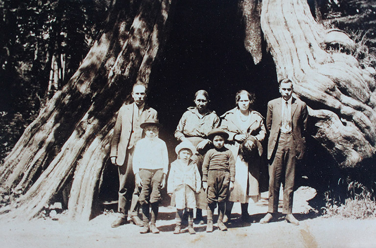 A family poses in front of the trunk of a very large tree. Four adults stand in the back, and three children stand in front.