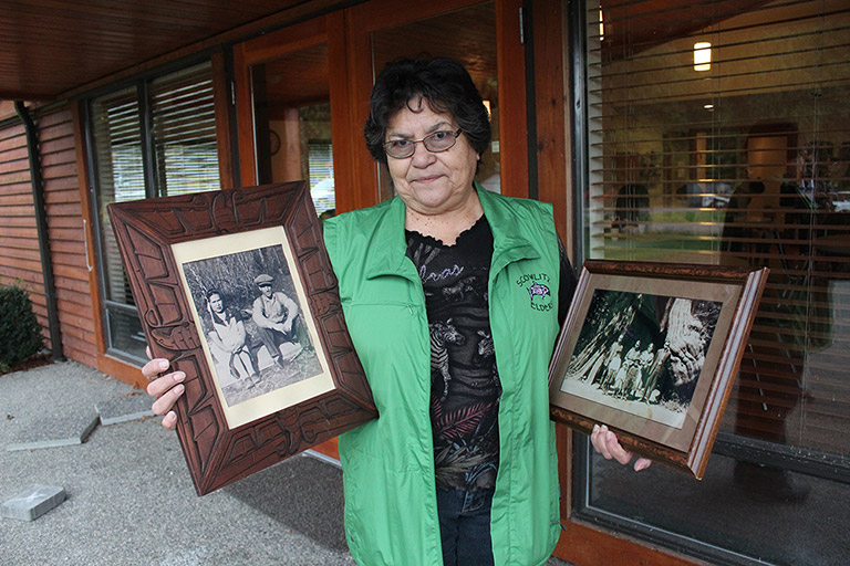 A woman stands beside a building, holding up a framed black and white photograph in each hand.