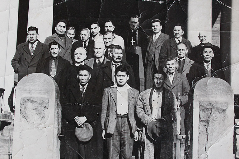 Une photo en noir et blanc d’un groupe de vingt hommes. Ils sont debout en trois rangées à l’extérieur d’un bâtiment et portent des tenues soignées.