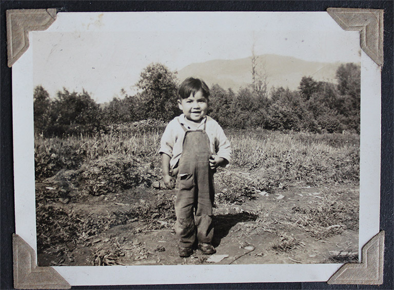A black and white photograph of a young boy standing in a field. The boy is wearing overalls and a long-sleeved white shirt.