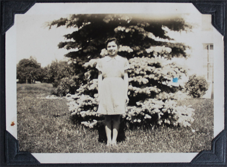 Une photo en noir et blanc d’une femme debout devant un arbre. Elle porte une robe à manches courtes.