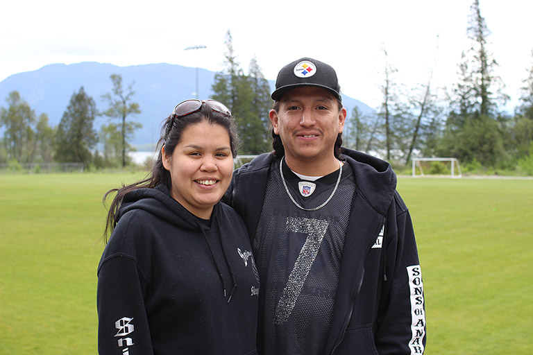 A woman and man stand next to each other in front of a soccer field. There are trees and mountains in the background.