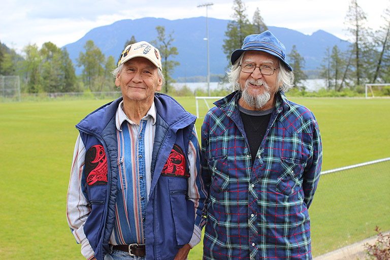 Two men stand in front of a soccer field. In the background there are trees, water, and mountains.