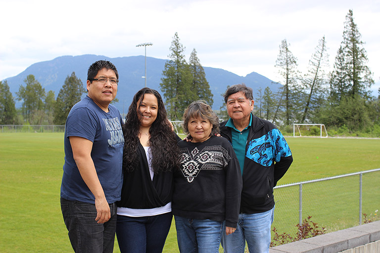 Two women stand between two men in front of a soccer field. In the background there are trees, water, and mountains.