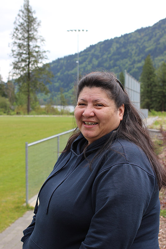 A woman stands in front of a soccer field. In the background there are trees, water, and mountains.
