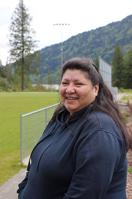 Une femme debout devant un terrain de soccer. À l’arrière-plan, il y a des arbres, de l’eau et une montagne.