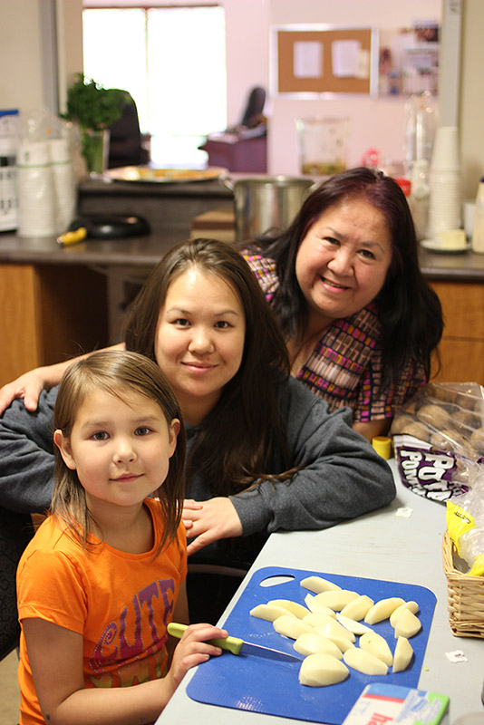 A girl, a young woman, and a woman are in a kitchen peeling potatoes. The woman has her arm around the young woman.