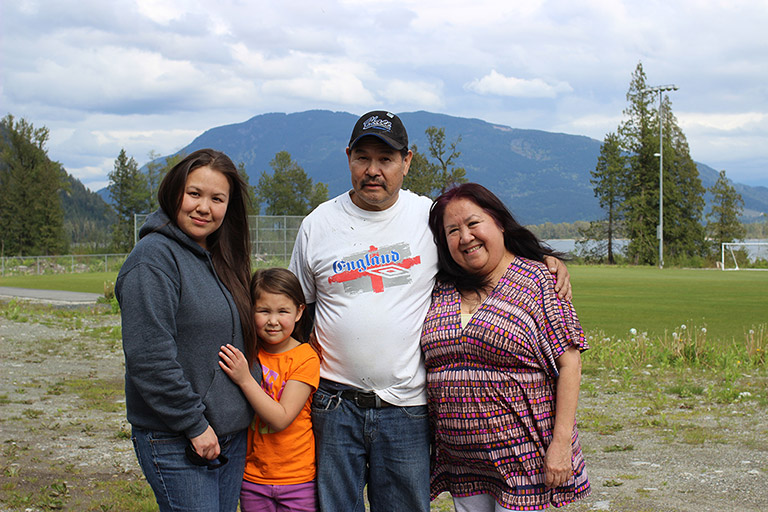 Two woman, a man, and a girl stand in front of a soccer field. In the background there are trees, water, and mountains. The young girl has her arms around the woman on the left.