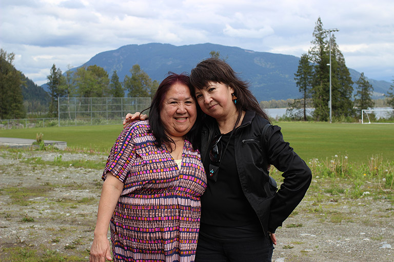 Two women stand in front of a soccer field with their arms around each other. In the background there are trees, water, and mountains.