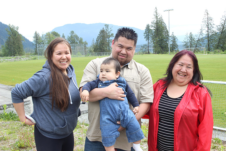 A man stands between two women in front of a soccer field. The man is holding a baby. In the background are some trees and mountains.