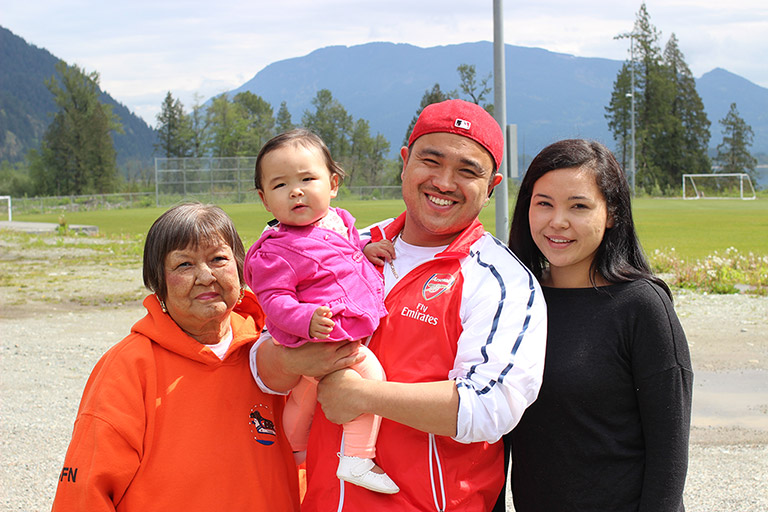 A man stands between two women in front of a soccer field. The man is holding a baby. In the background are some trees and mountains.