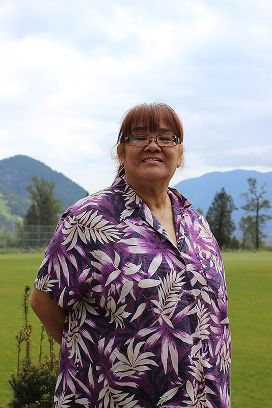 A woman stands in front of a soccer field. She has her hands behind her back. In the background are some trees and mountains.