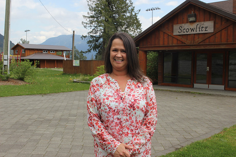 A woman stands in front of a building. Her hands are folded in front of her and she is smiling at the camera.