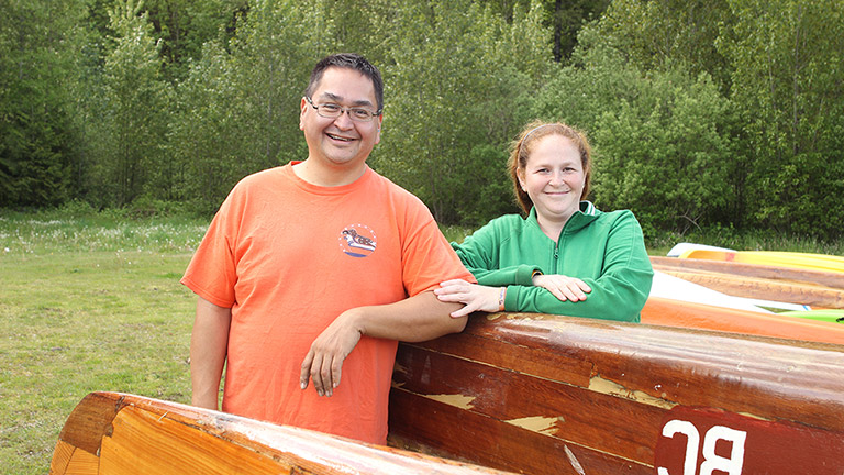 A man and woman stand outside beside a canoe, resting their arms on it. The woman is touching the man’s arm with her hand.