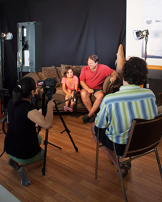 A man and a young girl sit on a couch as they are interviewed. A woman is kneeling in front of a camera on a tripod, while a male interviewer is sitting in a chair.