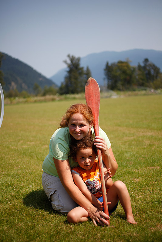 A woman kneels over a young boy in a field. The woman and boy hold a wooden paddle. In the background are some trees and mountains.