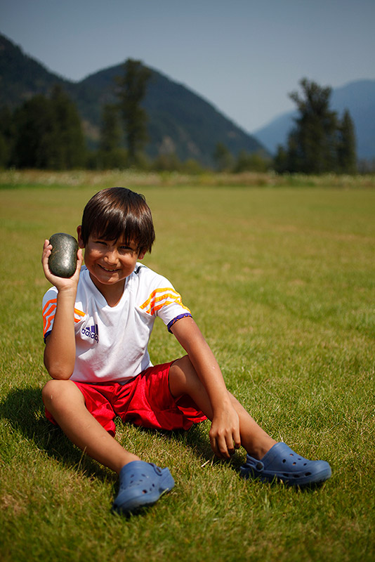 A boy sits in a field and smiles at the camera. In one of his hands he is holding up a grey object. In the background are some trees and mountains.