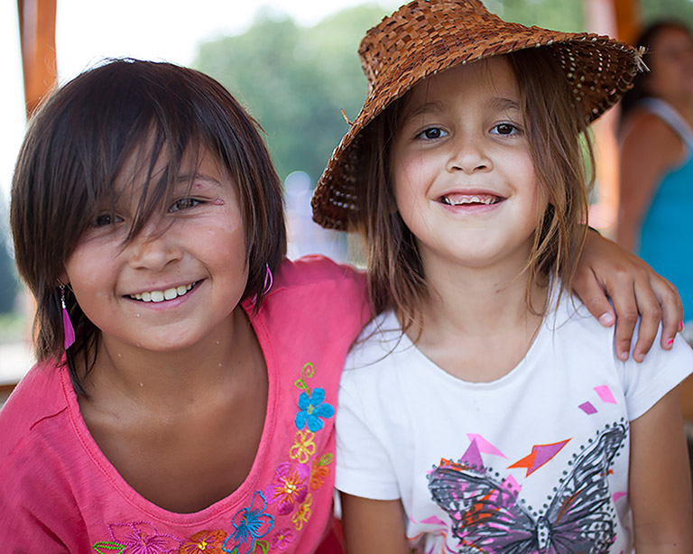 Two girls smile at the camera. The girl on the left is wearing a pink shirt and has her arm around the girl on the right. The girl on the right is wearing a white shirt and has a woven hat on her head.