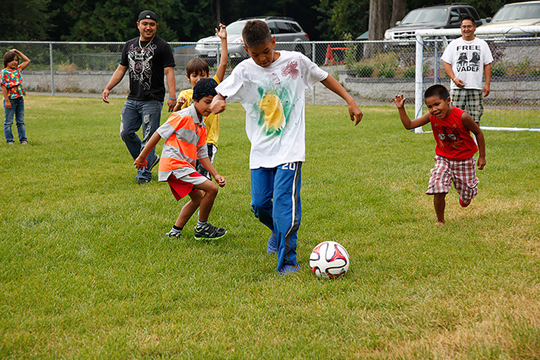 A group of men and children play soccer on a grass field. The children chase after a man dribbling with the ball.